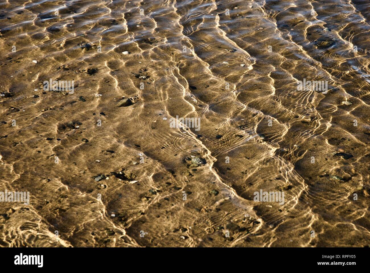 Sea water makes undulating abstract patterns and shapes in brilliant sunshine as it ripples and washes over a sandy beach Stock Photo