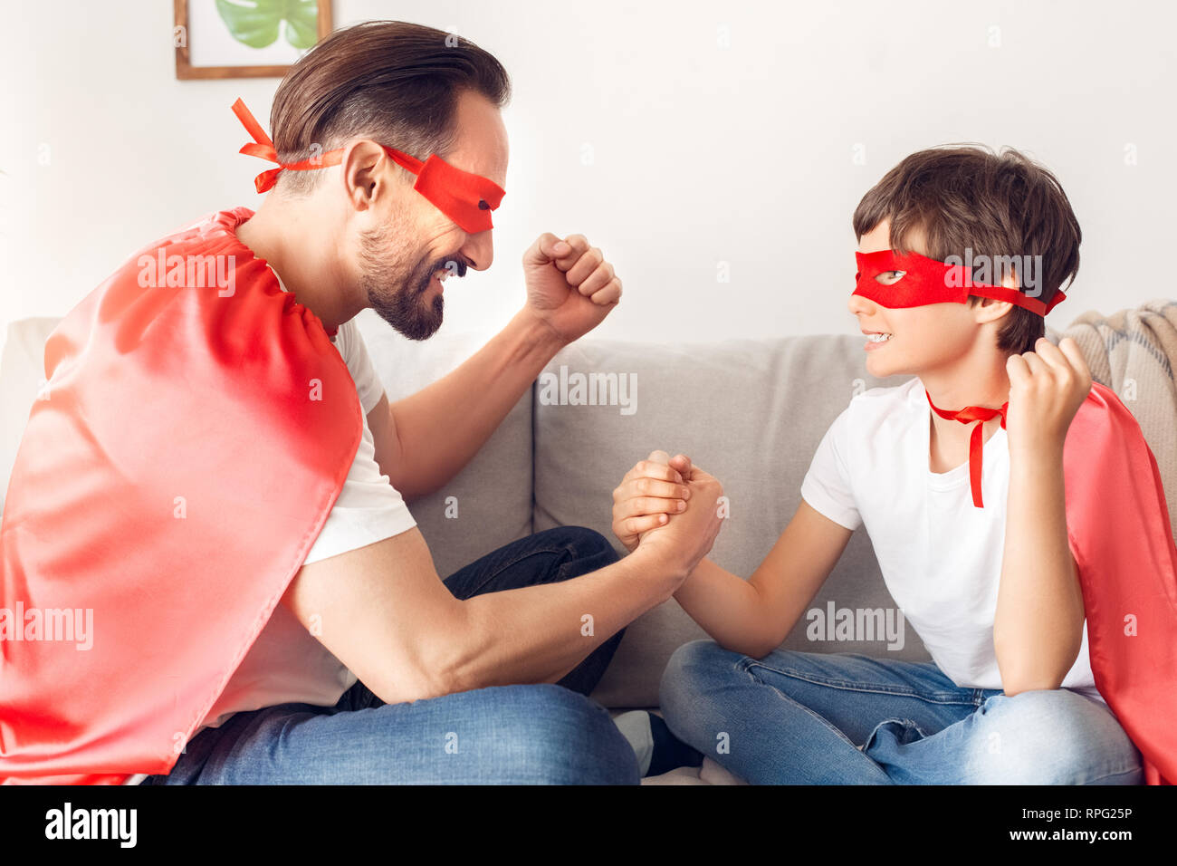 Father and son in superheroe costumes at home sitting on sofa holding hands looking at each other excited team spirit Stock Photo