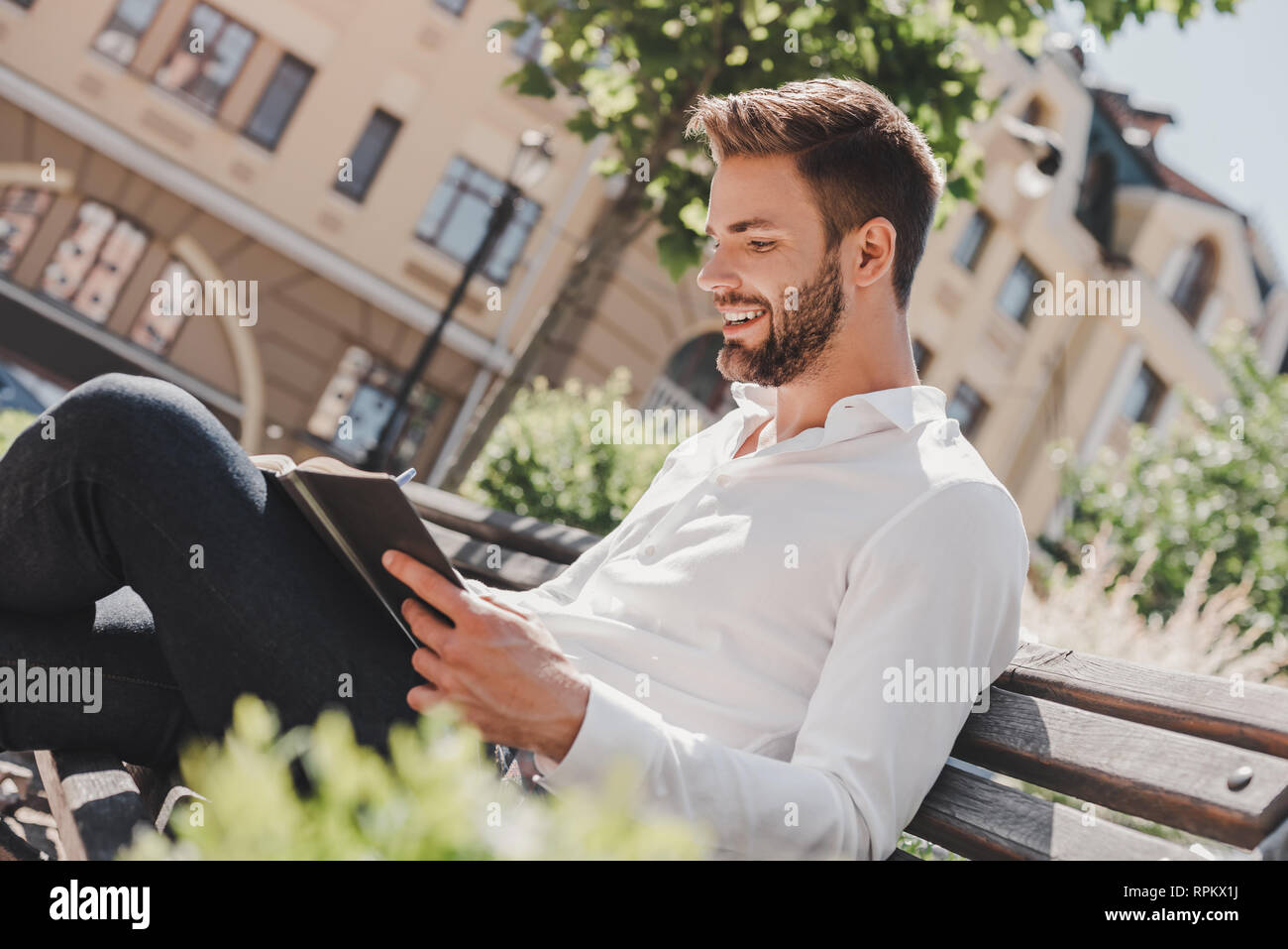 Close up of handsome young man sitting on a bench in the park and holding his planner. Bearded man dressed in a white shirt and jeans looking through his notes and smiling. Summertime, relax Stock Photo