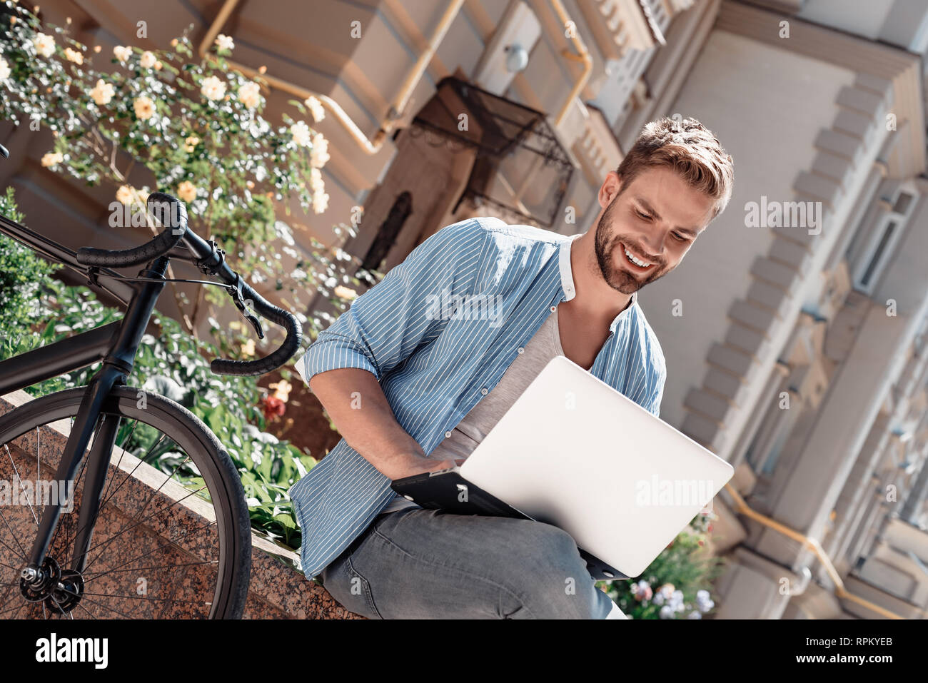 Portrait of handsome young man sitting in the park and holding his laptop. Bearded brown-haired man dressed in a blue striped shirt and jeans looking in his laptop and smiling. Summertime, relax Stock Photo
