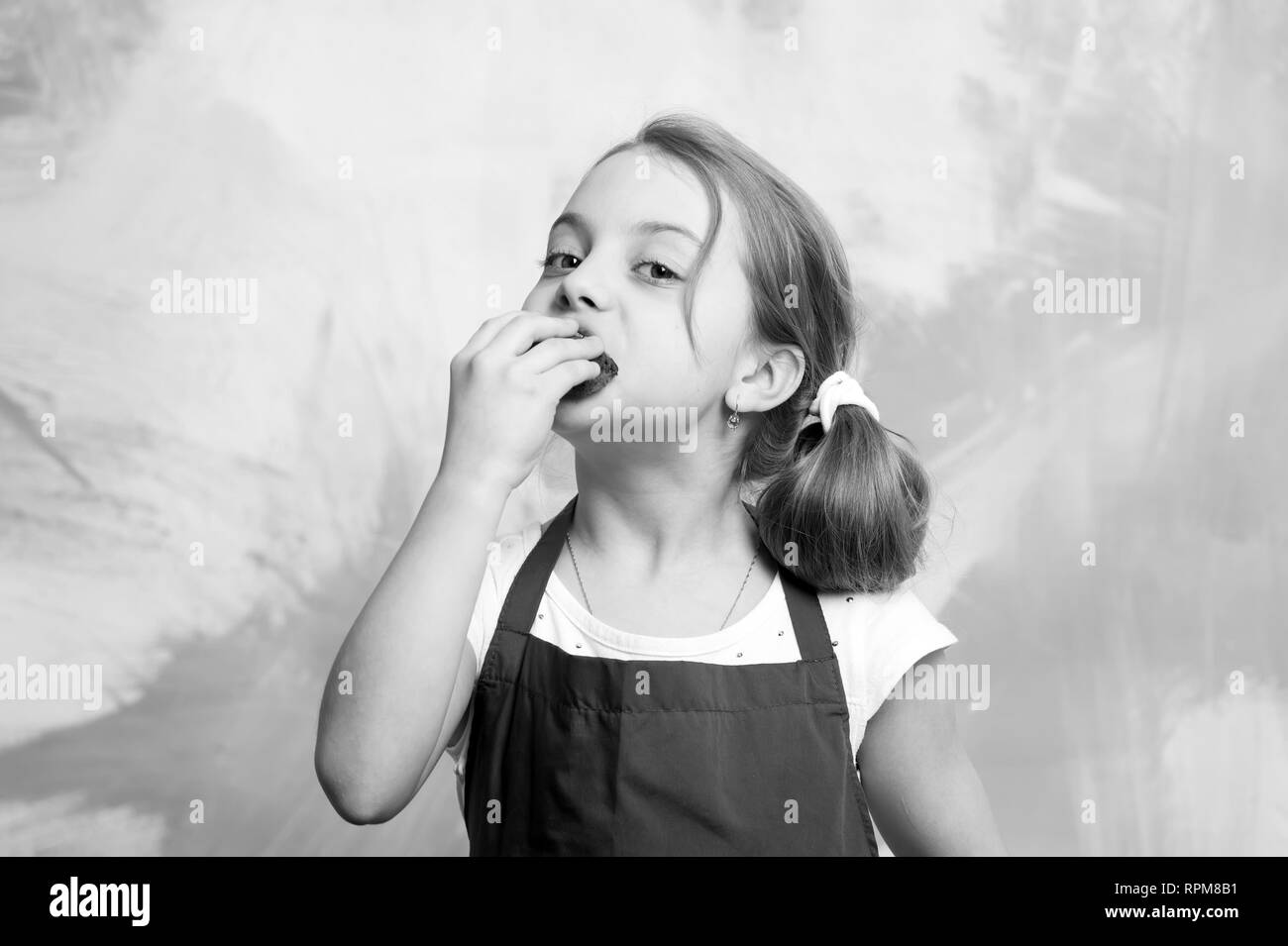 Child biting cake. Girl eating cupcake. Healthy food and diet concept. Baking and delicious bakery. Baby cook in red chef apron on colorful abstract wall. Stock Photo