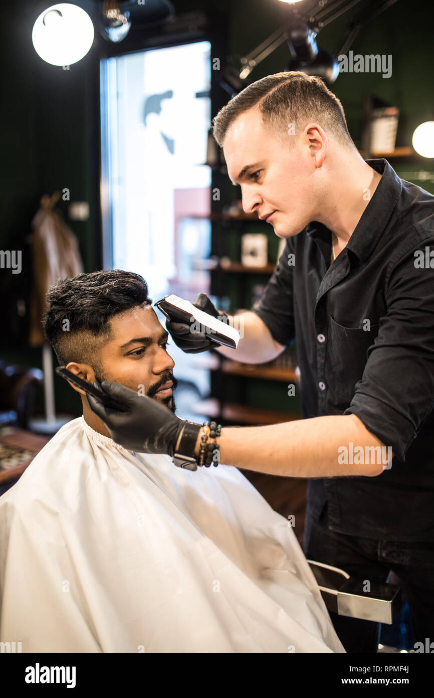 Professional styling. Close up side view of young bearded man getting haircut by hairdresser with electric razor at barbershop Stock Photo