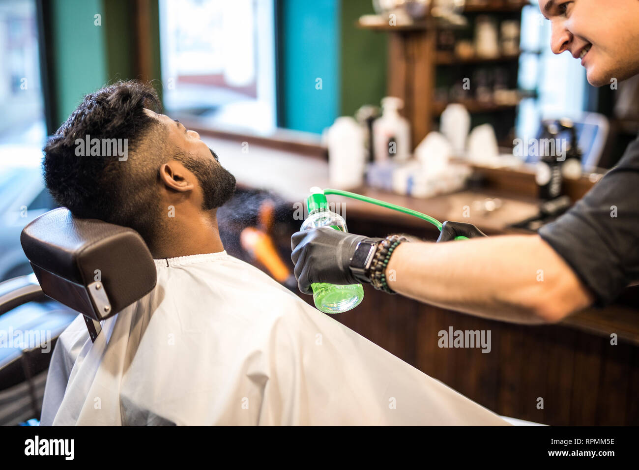 Young man sitting in barber chair combing and spraying hair of little kid with hands barber Stock Photo