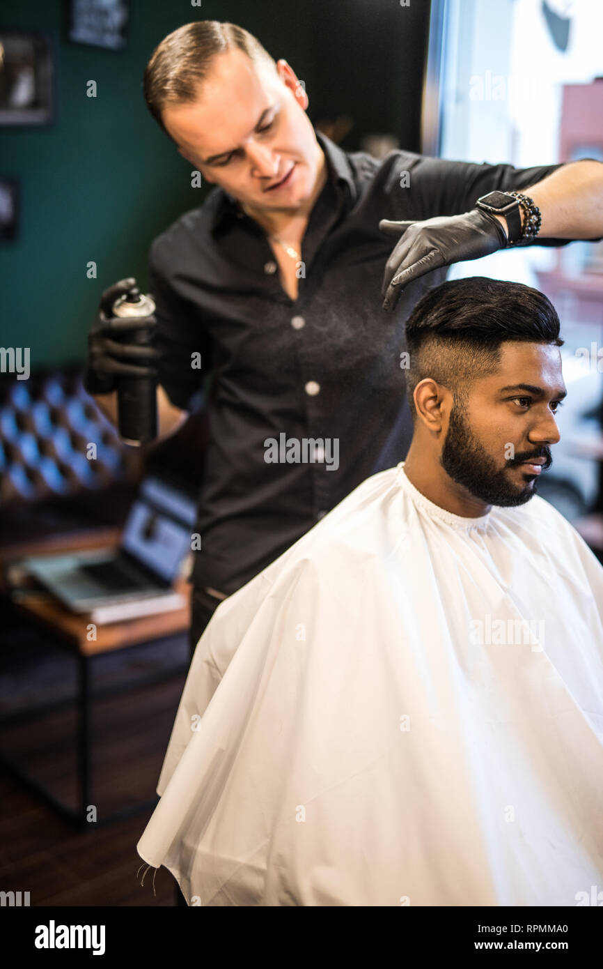 Barber combs his hair to young guy with a beard and mustache and sprinkles  hair on the spray gun. Barber Shop Stock Photo - Alamy