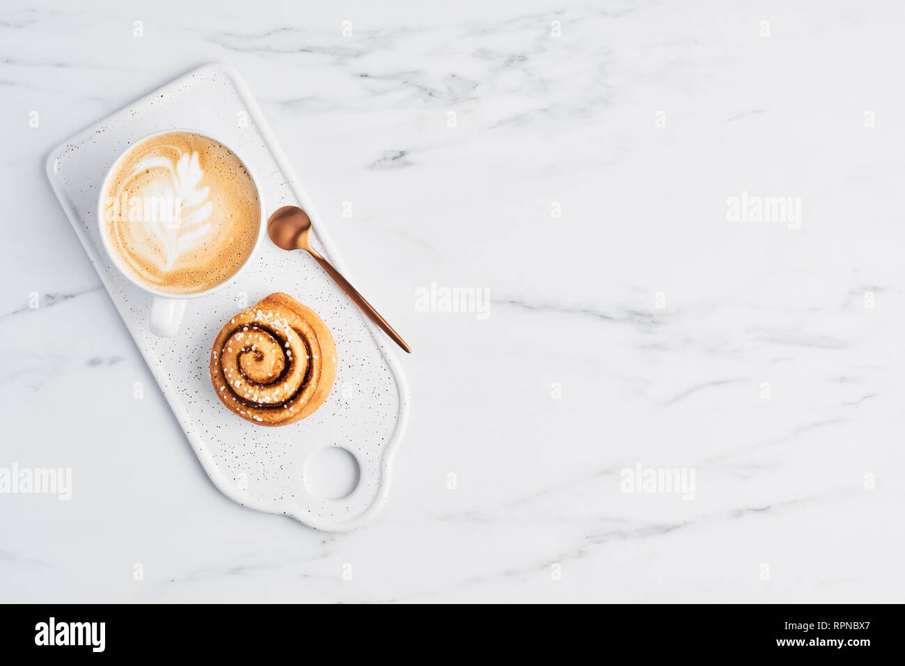 Freshly baked cinnamon bun with spices and cocoa filling and coffee or cappuccino with latte art on white serving plate over white marble background.  Stock Photo