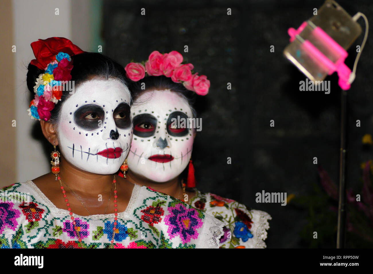 Mexican teenagers, young women, with traditional outfits, flowers and skull make up, are using a pink selfie stick during celebration of Day of the de Stock Photo