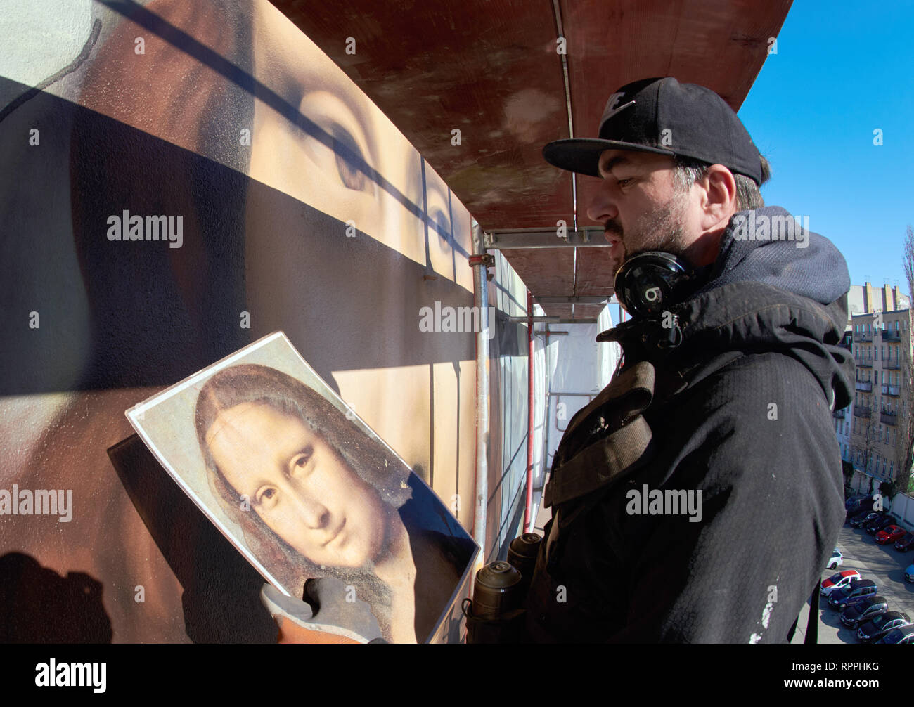 22 February 2019, Berlin: The artist Tank stands high up on the scaffolding, opposite the East Side Gallery, and looks at his model of Da Vinci's Mona Lisa to transfer the work from the Louvre in Paris as faithfully as possible to the windowless façade of the East Side Hotel. Together with the artists Tank, Weisse Seite and FIX77, the Berlin artist collective Die Dixons has painted an oversized Mona Lisa on the firewall of a hotel in Mühlenstraße opposite the East Side Gallery. With an area of around 170 square metres, it is the largest exact Mona Lisa with the same shape in the world. Photo: Stock Photo