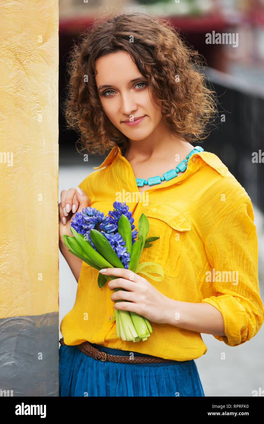 Happy young fashion woman with bouquet of flowers walking in city street Stylish female model wearing yellow shirt and blue skirt Stock Photo