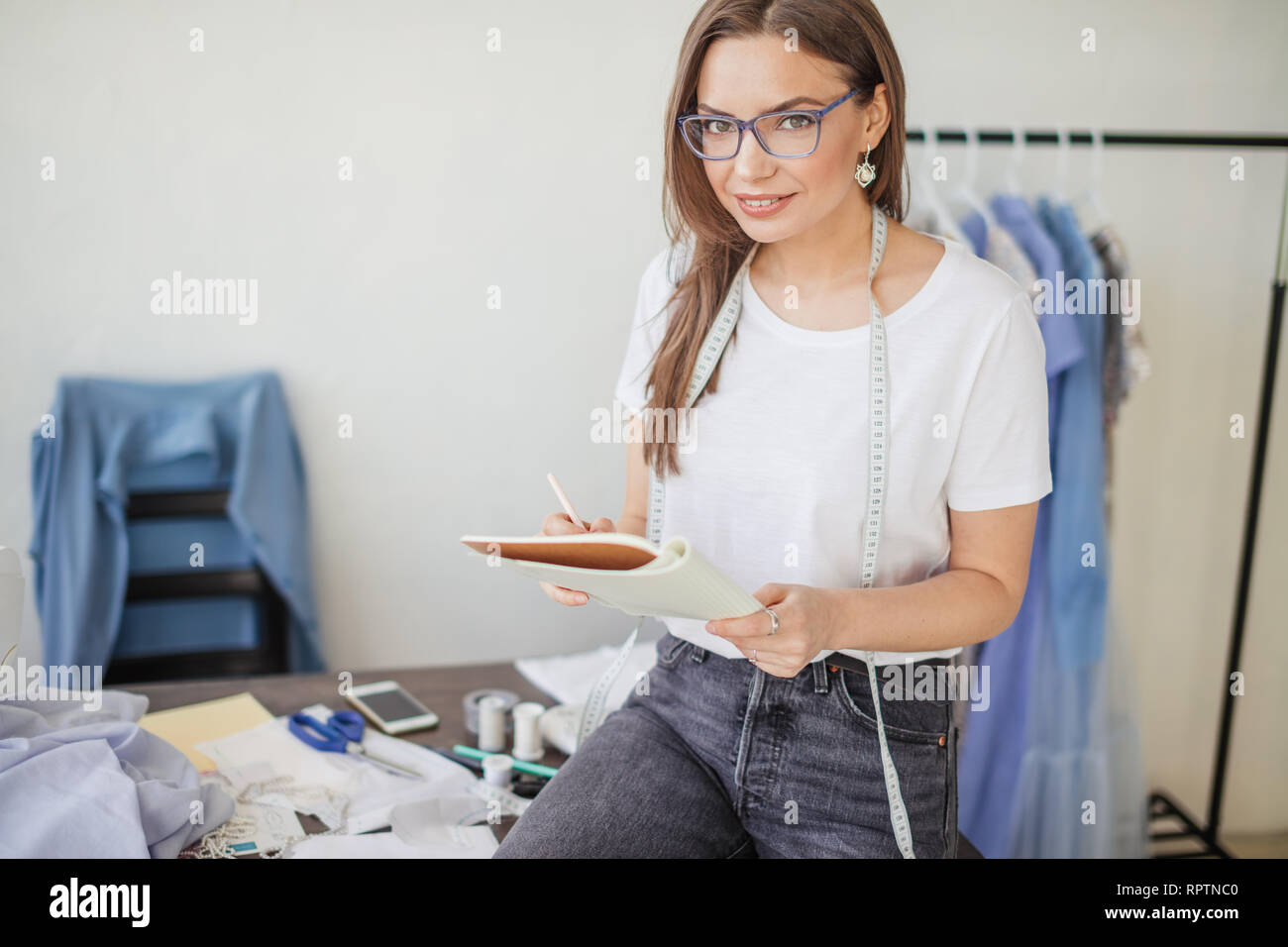 Young hard working seamstress with long blonde hair and spectacles sitting at her studio workplace and creating new images of her own design clothes Stock Photo