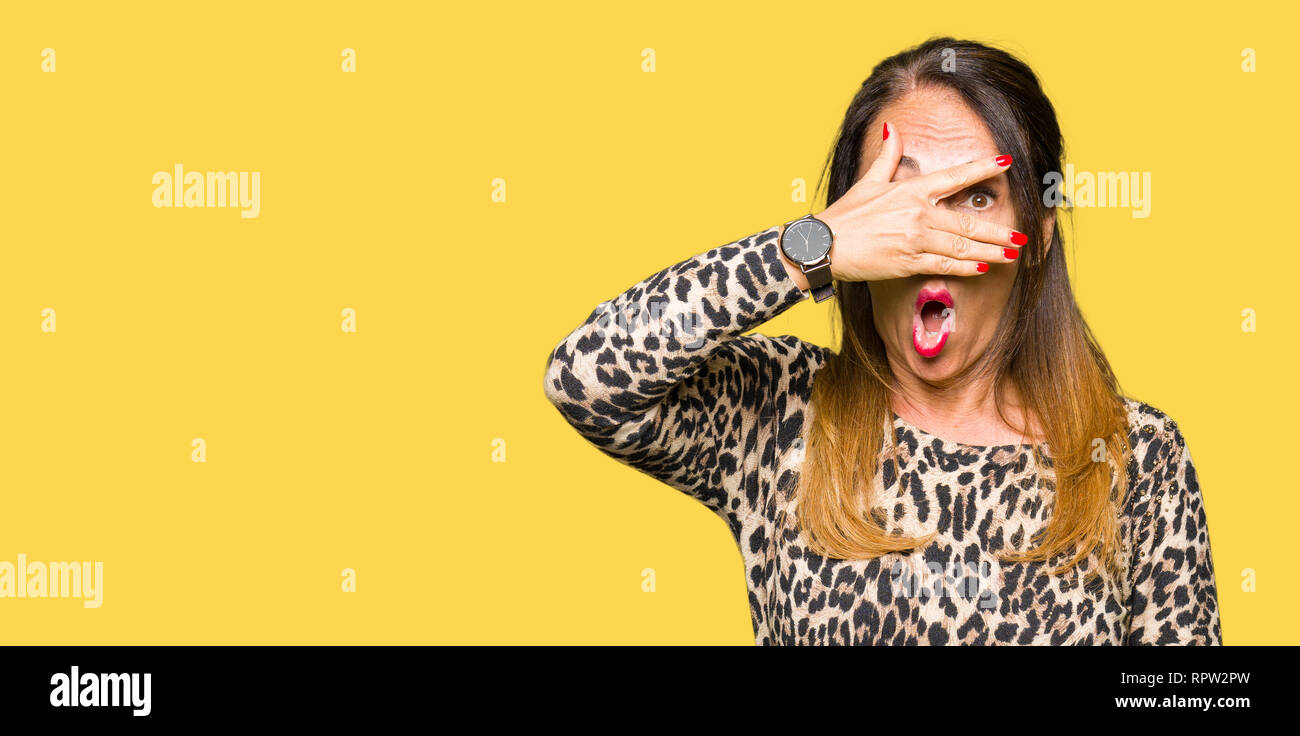 Beautiful middle age woman wearing leopard animal print dress peeking in shock covering face and eyes with hand, looking through fingers with embarras Stock Photo
