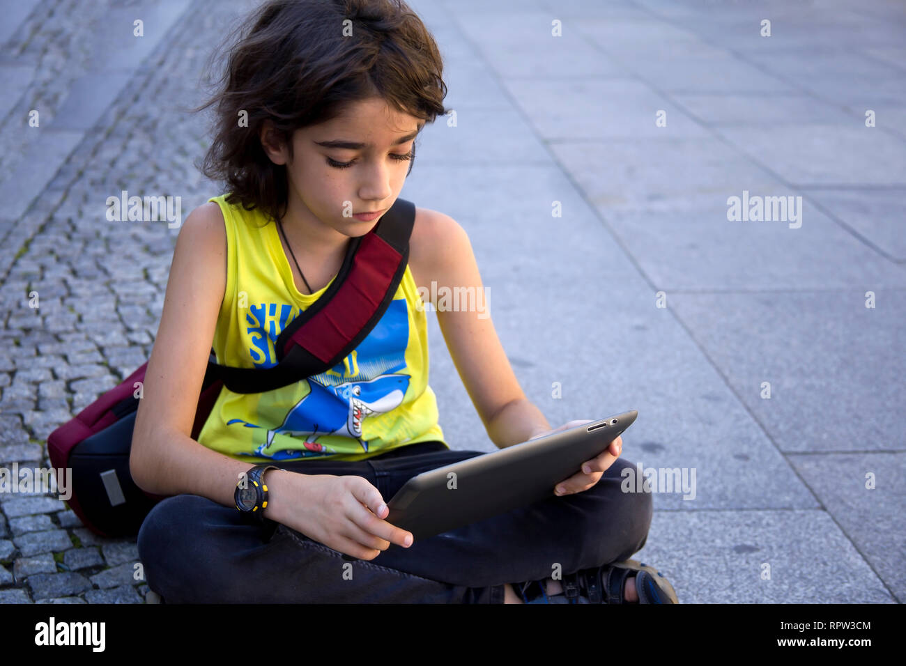 Teenage boy with long brown hair sitting outside on concrete using a tablet computer, looking down while sitting with crossed legs. Stock Photo