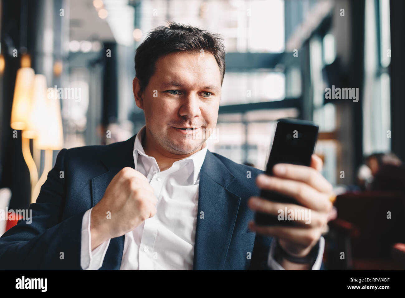 Excited young businessman sitting in a bar and holding mobile phone, raising a hand in celebration after receiving the news of making money on an inve Stock Photo