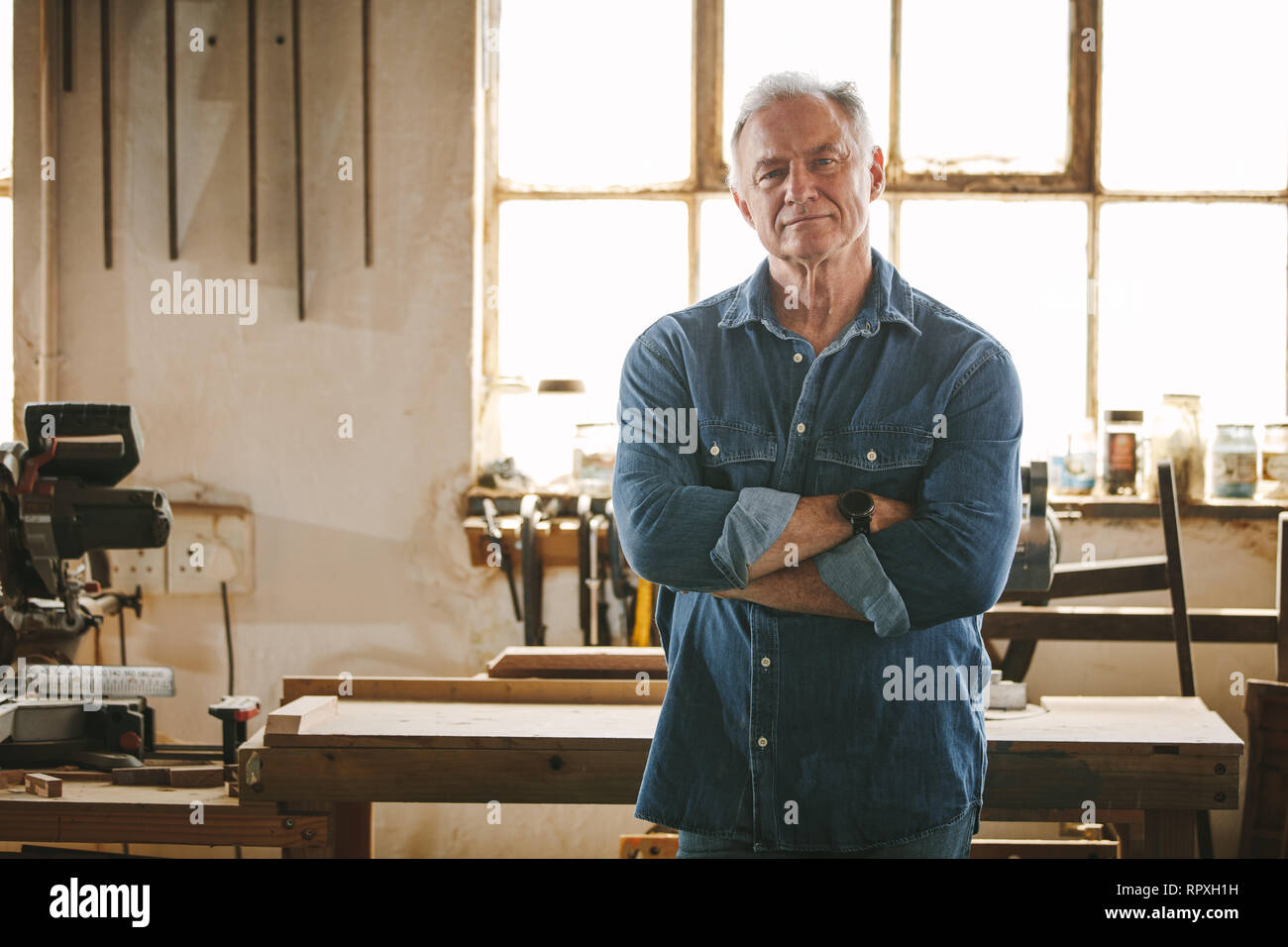 Portrait of confident mature male carpenter standing in his carpentry workshop. Senior caucasian man looking at camera while standing in a carpentry. Stock Photo