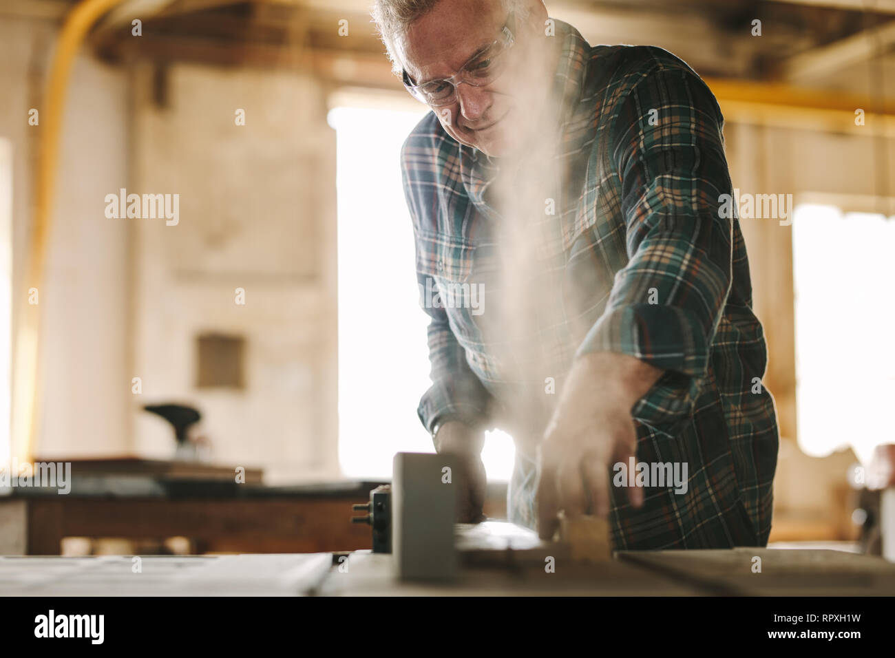 Mature male carpenter working on table saw machine in carpentry workshop. Senior man cutting wood on table saw machine. Stock Photo