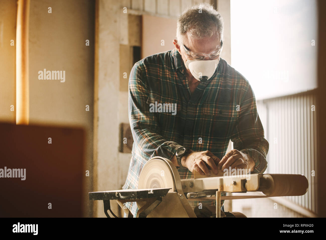 Senior carpenter sanding a wood with belt sander in carpentry workshop. Male carpenter with face mask working on belt sander machine. Stock Photo