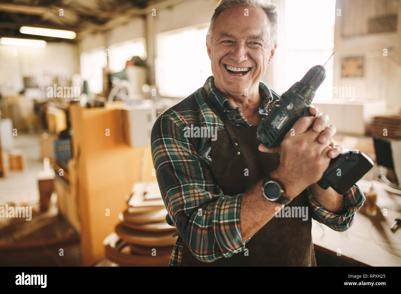 Smiling senior carpenter holding drill machine against workshop. Mature male worker smiling confidently to the camera holding drilling machine at his  Stock Photo