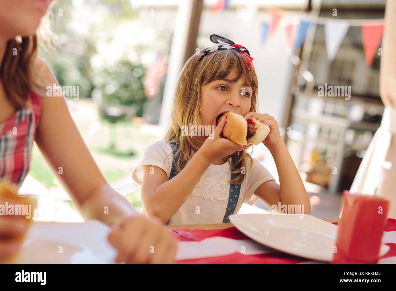 Girl eating a hot dog sitting with her family. Little girl enjoying a hotdog at a restaurant. Stock Photo