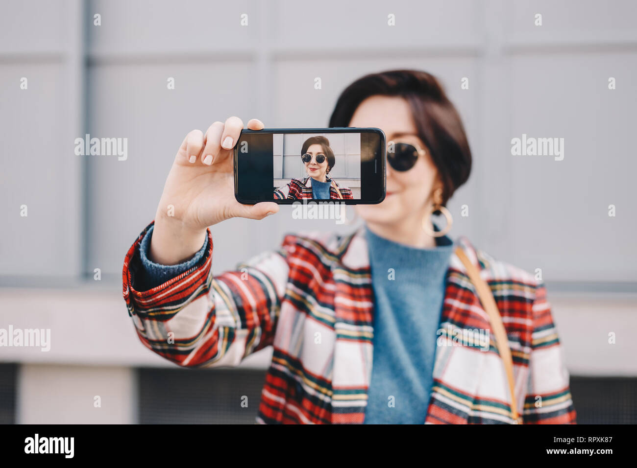 Close up portrait of pretty and modern young female influencer taking a selfie with her mobile phone, wearing fancy clothes and posing for followers o Stock Photo