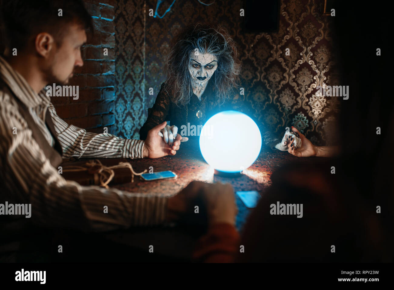 Scary witch reads a magic spell over a crystal ball, young man and woman on spiritual seance. Stock Photo