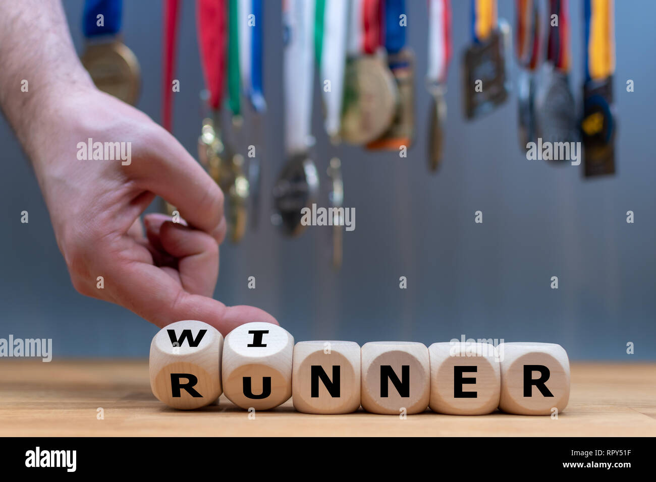 Hand turns a dice and changes the word 'RUNNER' to 'WINNER' in front of a background full of medals. Stock Photo