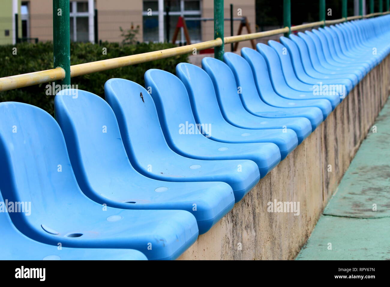 Row of new blue plastic seats mounted on top of concrete bleachers with metal fence and garden vegetation in background next to basketball court Stock Photo