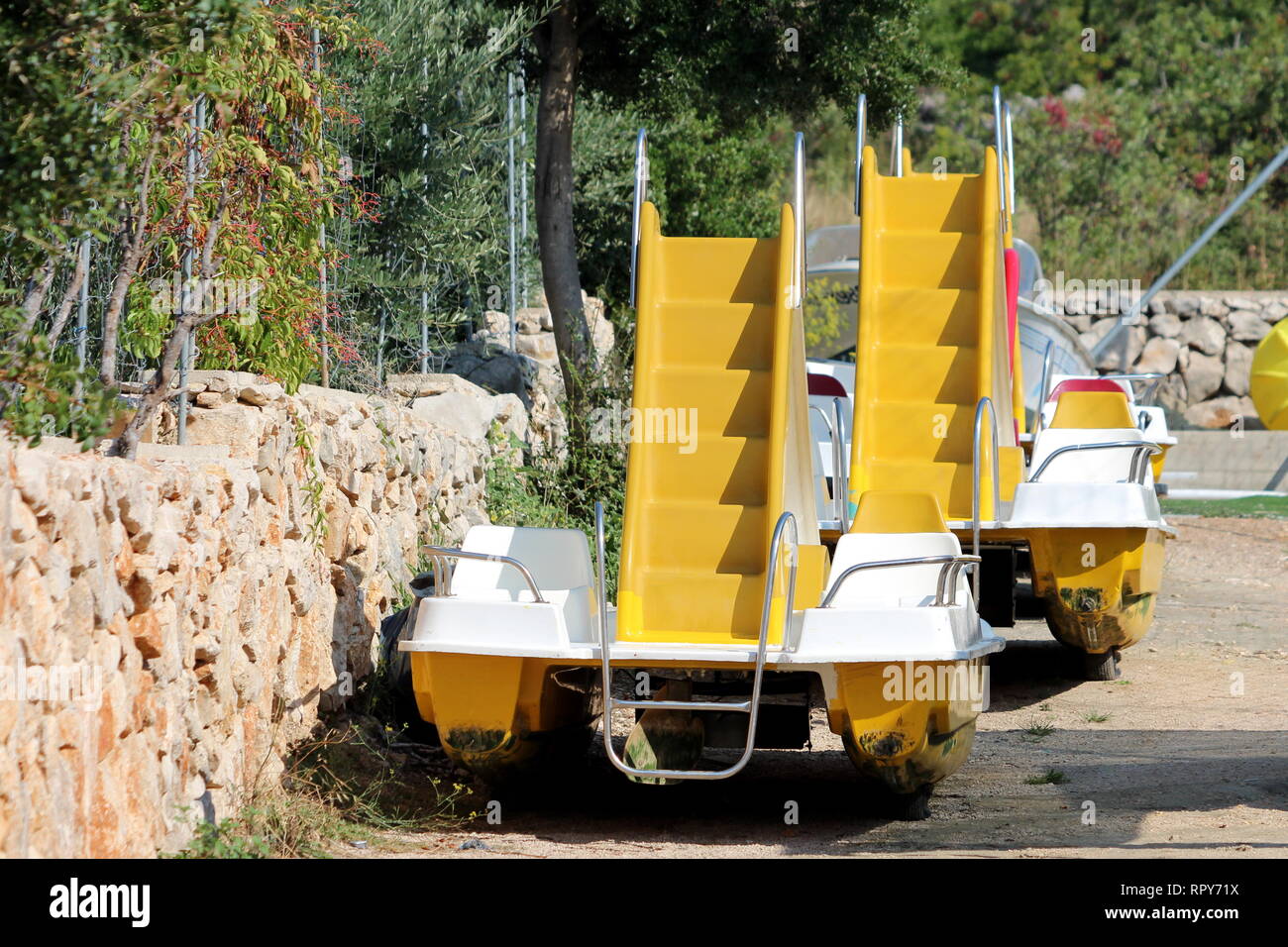 Two yellow paddle boats with plastic steps and slides in middle taken out of water for cleaning left in a row next to traditional stone wall Stock Photo