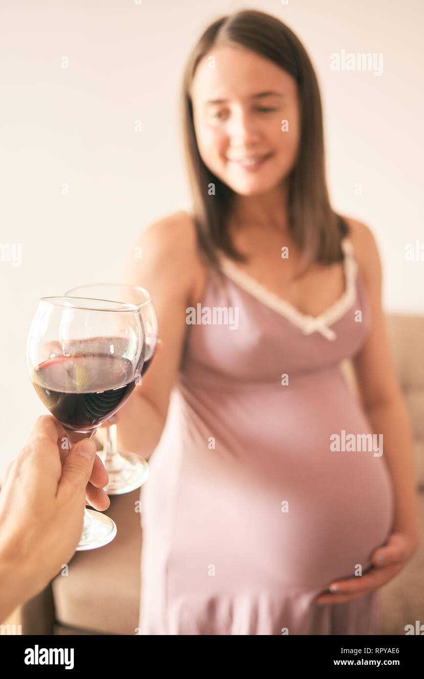 Front view of smiling pregnant woman standing at home and cheering with friend. Young future mother wearing nighty drinking alcohol. Concept of harmfu Stock Photo