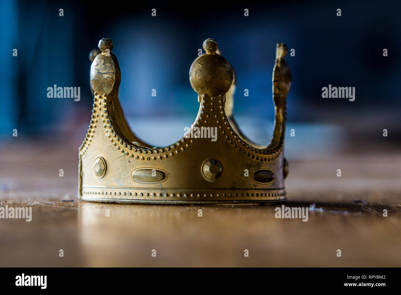 Close up of an isolated  worn out plastic gold-colored  crown which lies on a worn out wooden table with a blue background. Stock Photo
