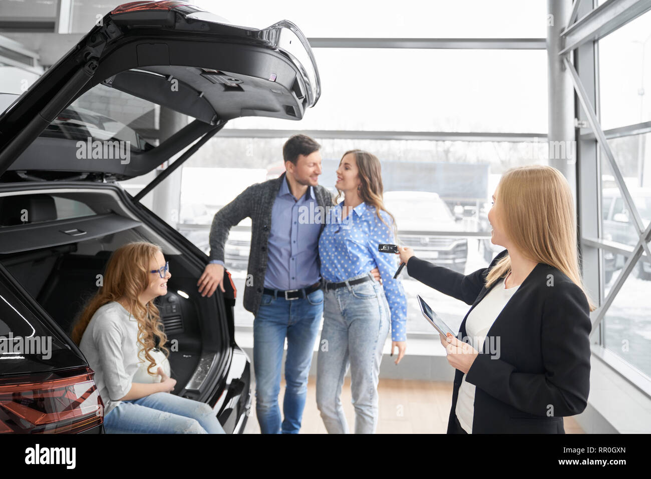 Manager of car dealership working with clients, holding folder and giving car keys to buyers of vehicle. Couple standing, posing, daughter sitting in car trunk, smiling. Stock Photo