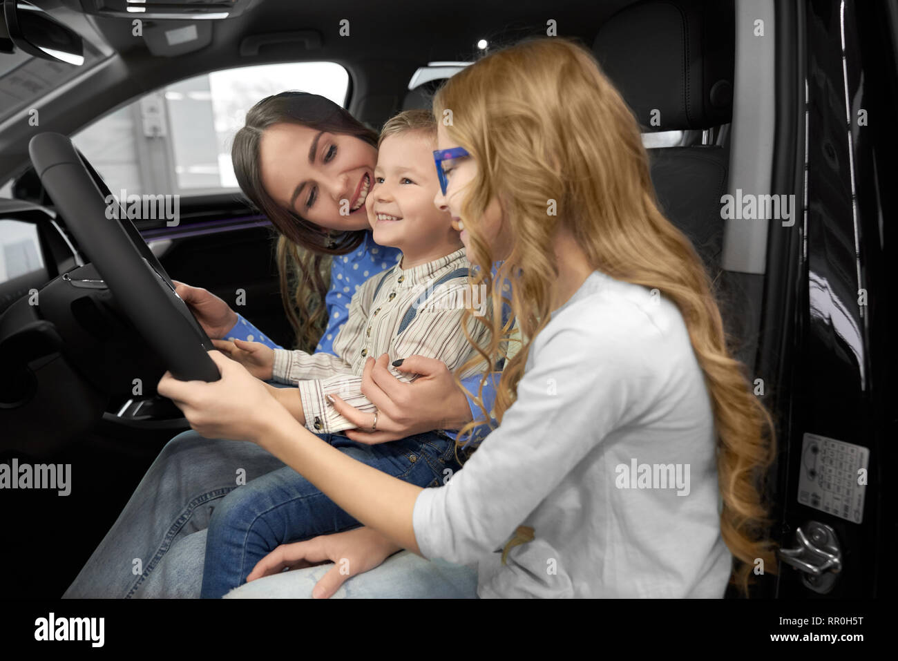 Mother sitting with children in car cabin of new automobile, observing and testing auto. Woman holding cute, happy son on knees, beautiful daughter holding hand on steering wheel. Stock Photo