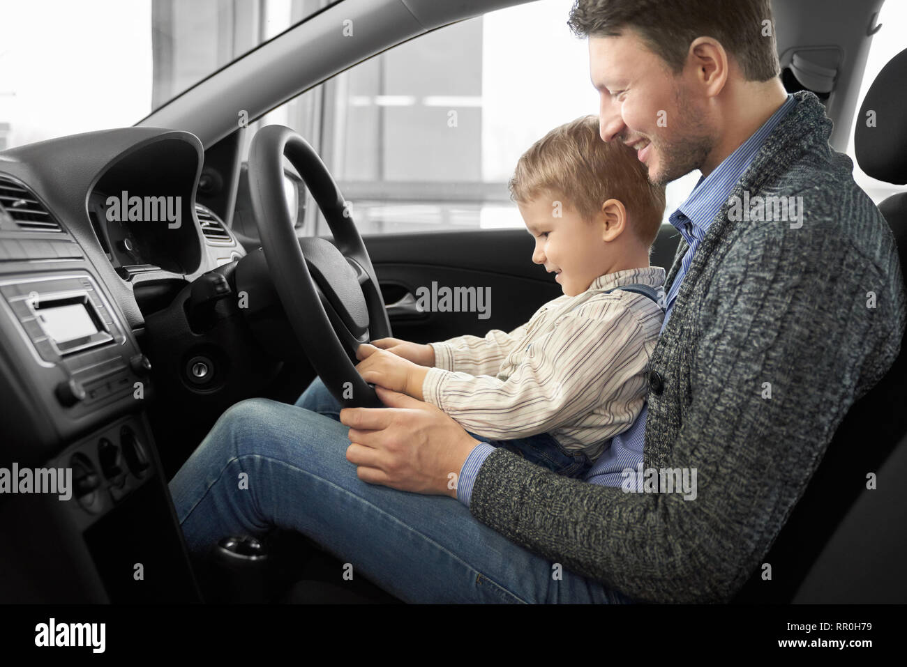 Father holding son on knees, man sitting in driver's seat of new expensive car. Parent showing to kid car cabin, steering wheel. Happy family observing automobile, smiling. Stock Photo