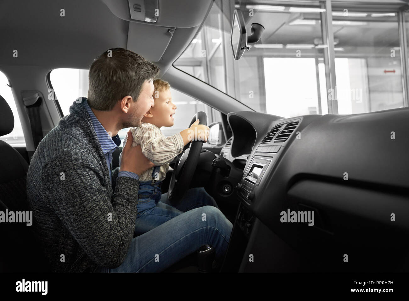 Happy father and son observing and testing new car in dealership. Parent holding kid on knees, sitting in driver's seat, car cabin. Little boy holding hands on steering wheel, smiling. Stock Photo