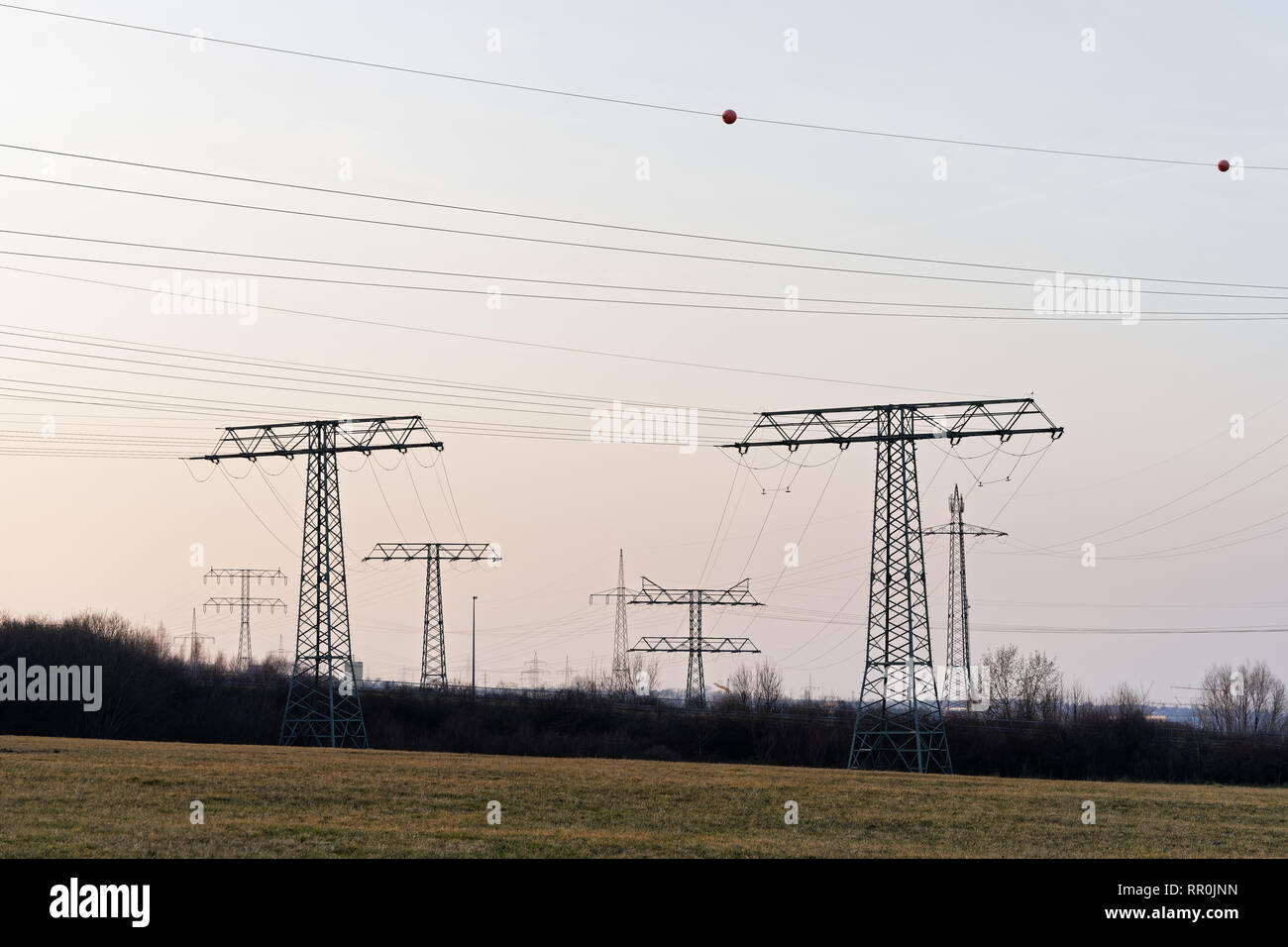 Large collection of electricity pylons in different types with cables, they stand staggered as silhouettes in front of a slightly colored evening sky  Stock Photo