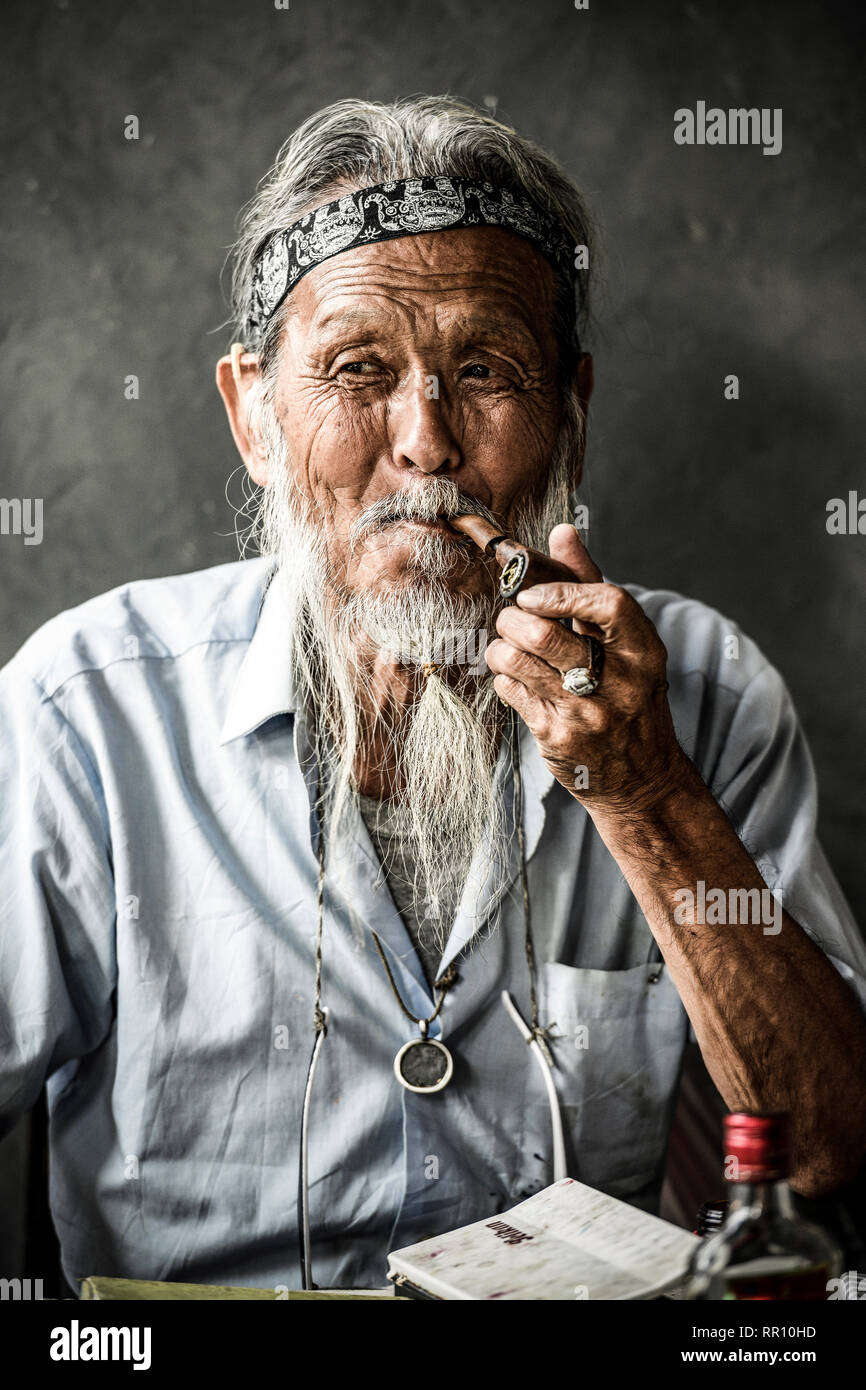 Portrait of an elderly Japanese man smoking a pipe while on the table there is a bottle of rum and painting tools. Osaka, Japan. Stock Photo