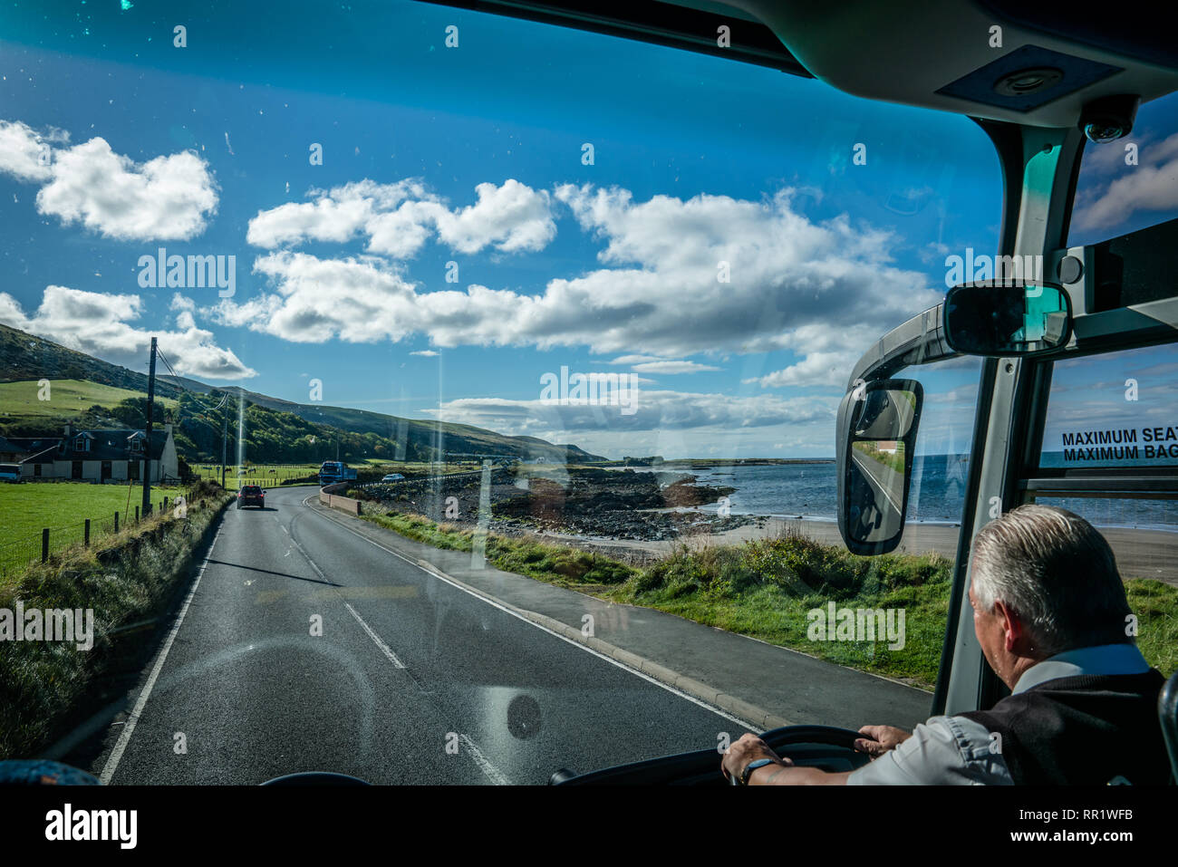Looking out the front window of a bus.  Seeing the bus driver and the countryside of Scotland. Green fields and ocean on both sides. Stock Photo