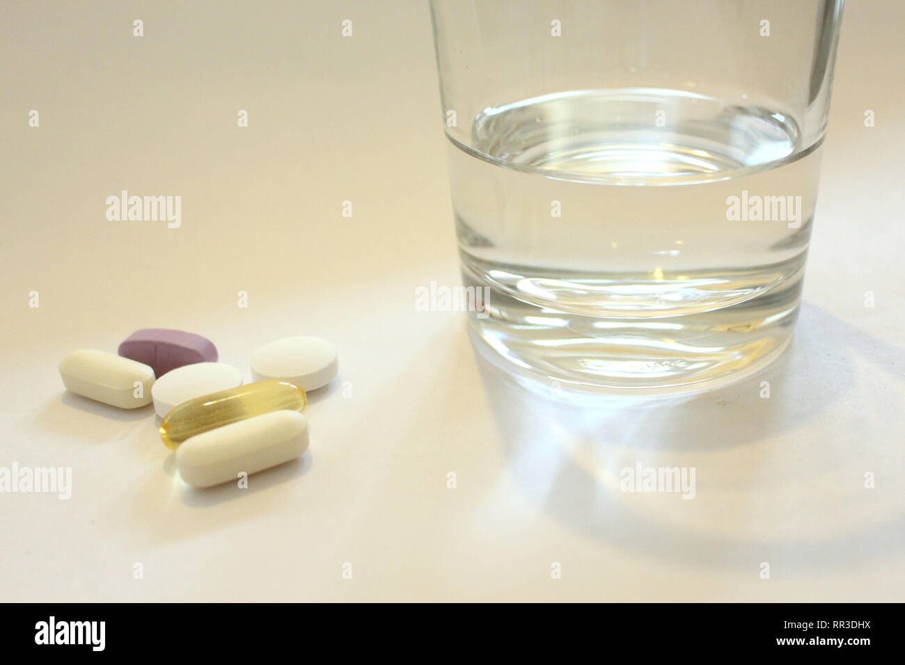 A photograph of a handful of various pills on a white background next to a glass of water. Concept of taking medication or supplements. Stock Photo