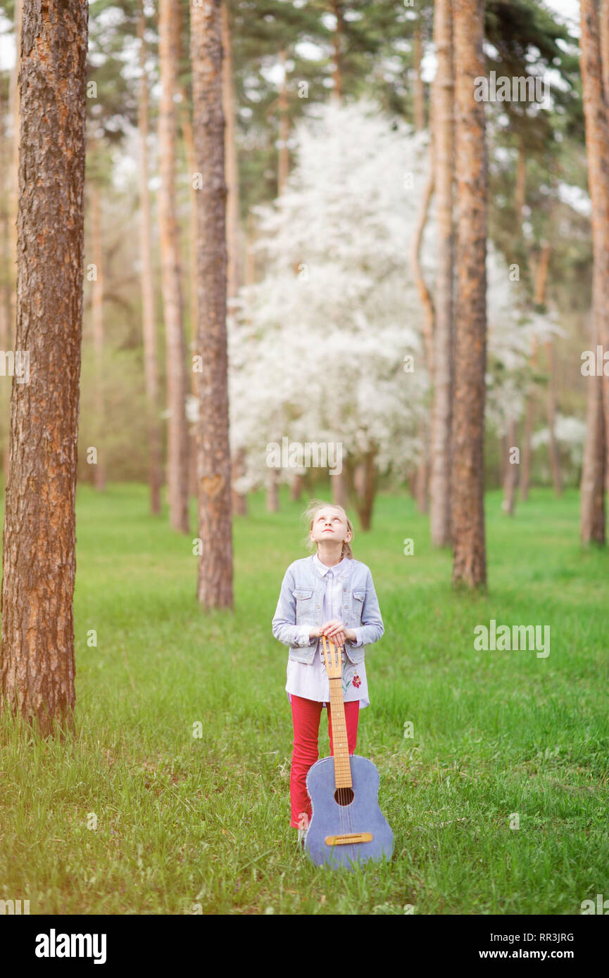 beautiful little girl with vintage guitar getting inspired standing among high trees in spring park Stock Photo
