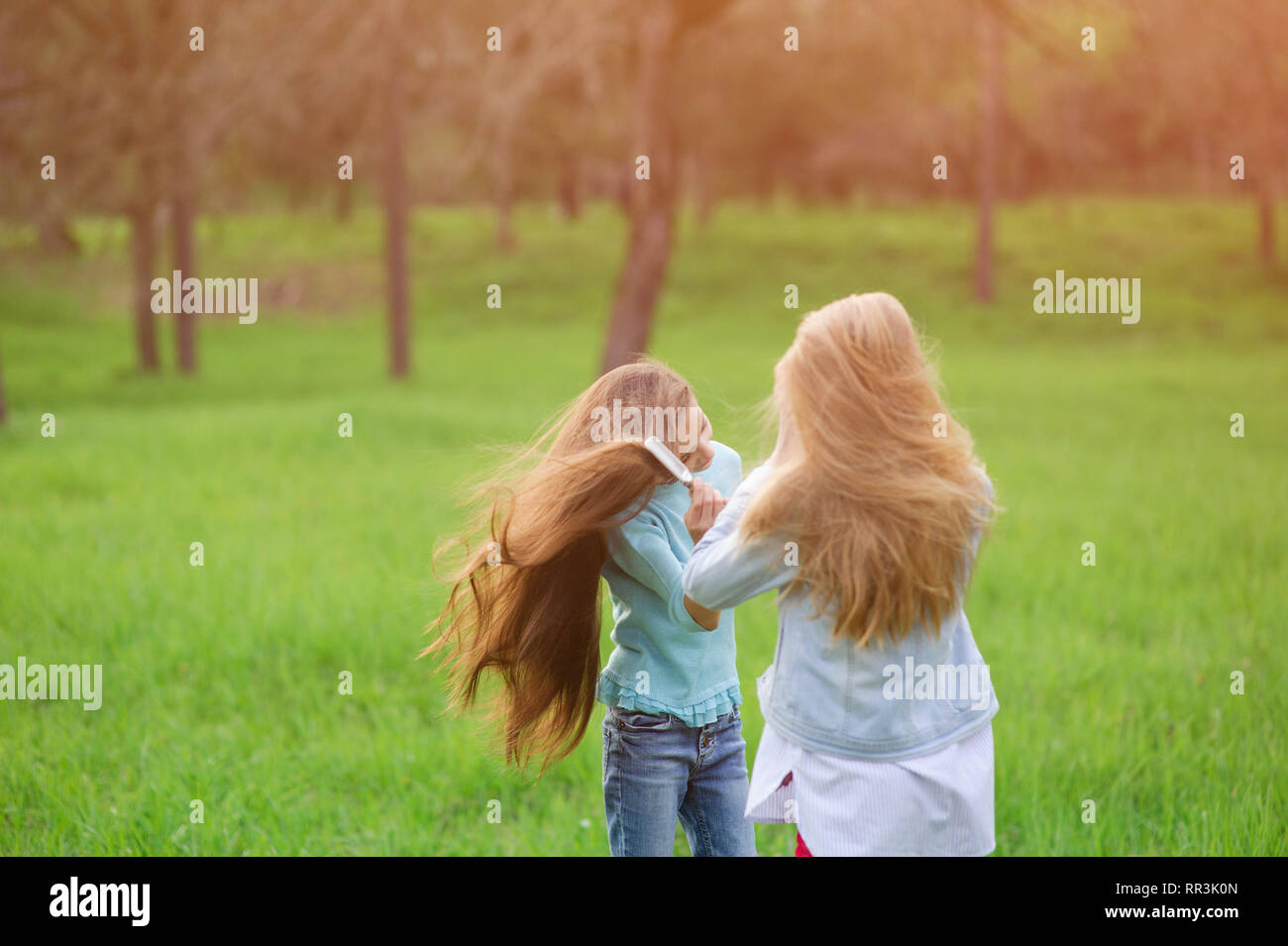 two little beautiful brown haired and blond girls comb hair with hairbrush outdoors in park Stock Photo