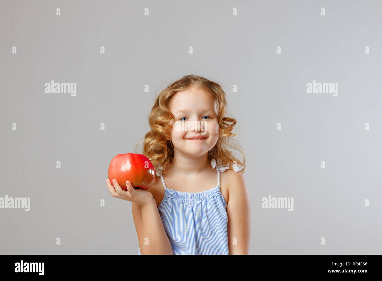 Happy little girl child holding a red apple. Healthy food. Gray background, studio, portrait Stock Photo