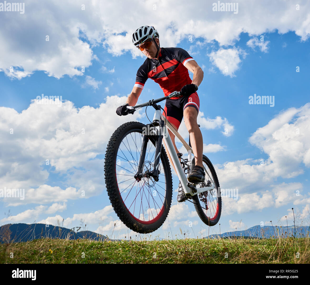 Young athletic sportsman biker in professional sportswear flying in air on his bike on bright blue sky with white clouds and distant hills background Active lifestyle and extreme sport concept. Stock Photo