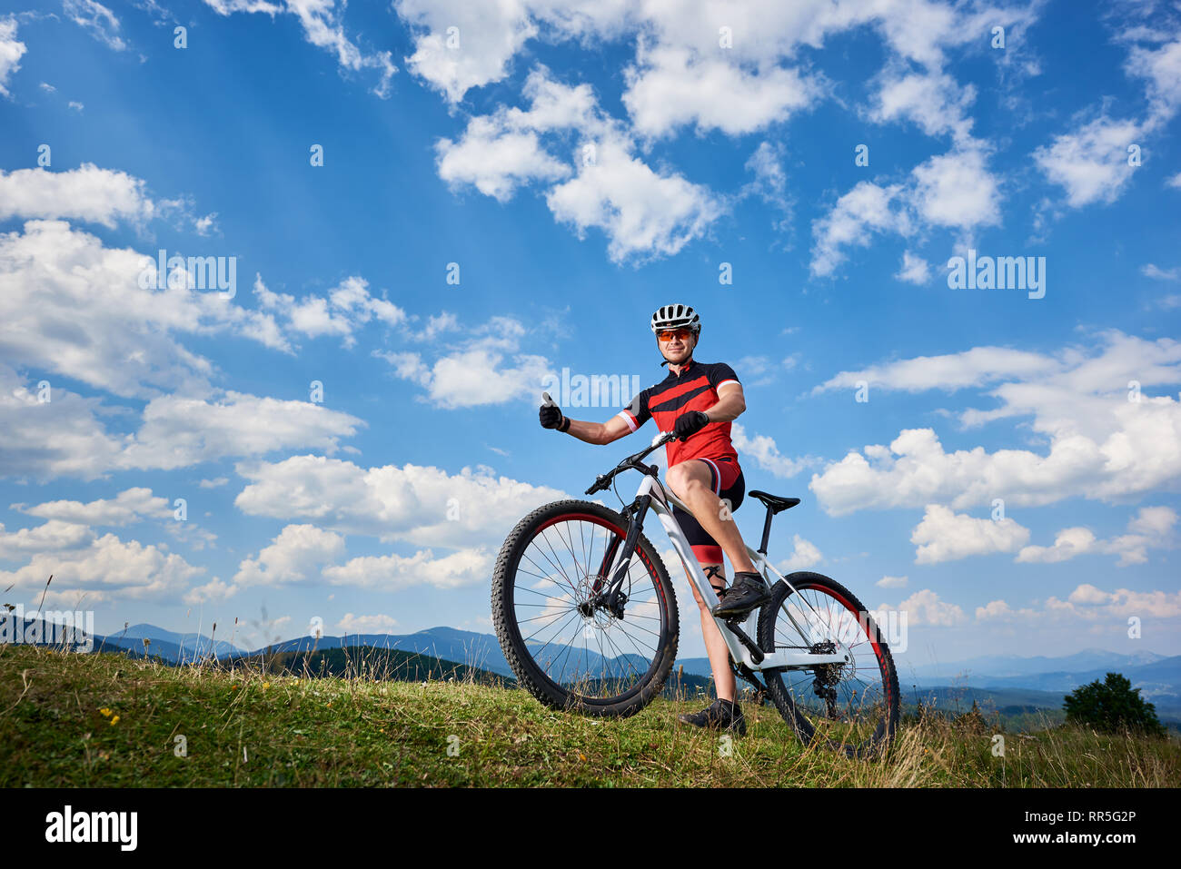 Smiling professional cyclist in sportswear and helmet standing with cross  country bike on top of hill,