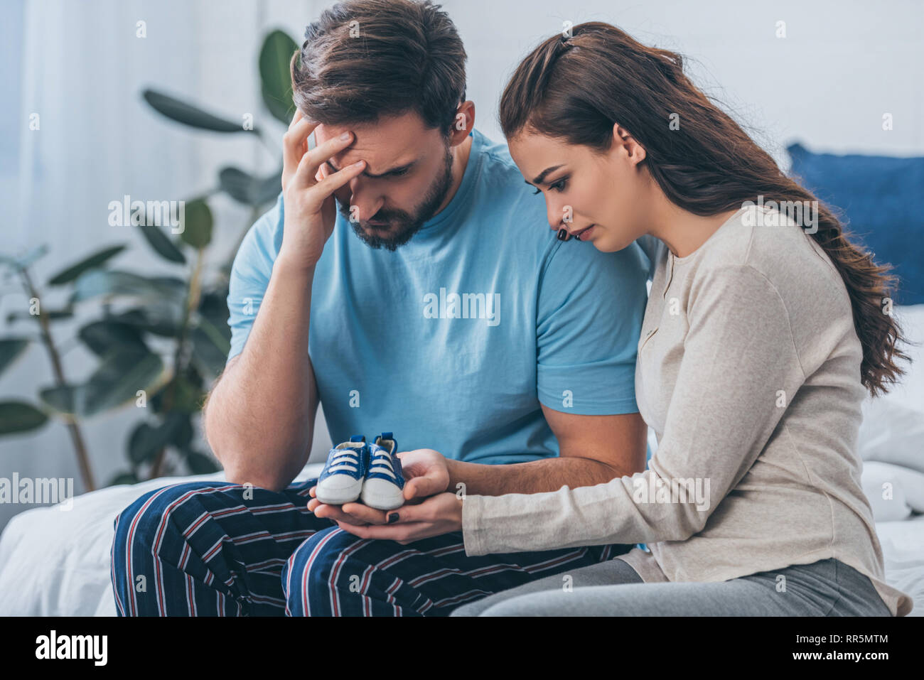 selective focus of grieving parents sitting on bed and holding baby shoes at home Stock Photo