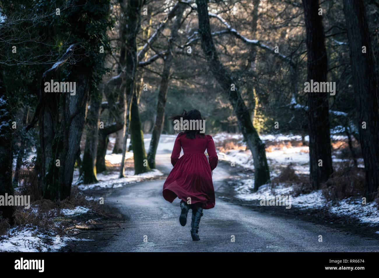 a young woman in a red dress on a small street in the middle of a wintery forest with snow Stock Photo