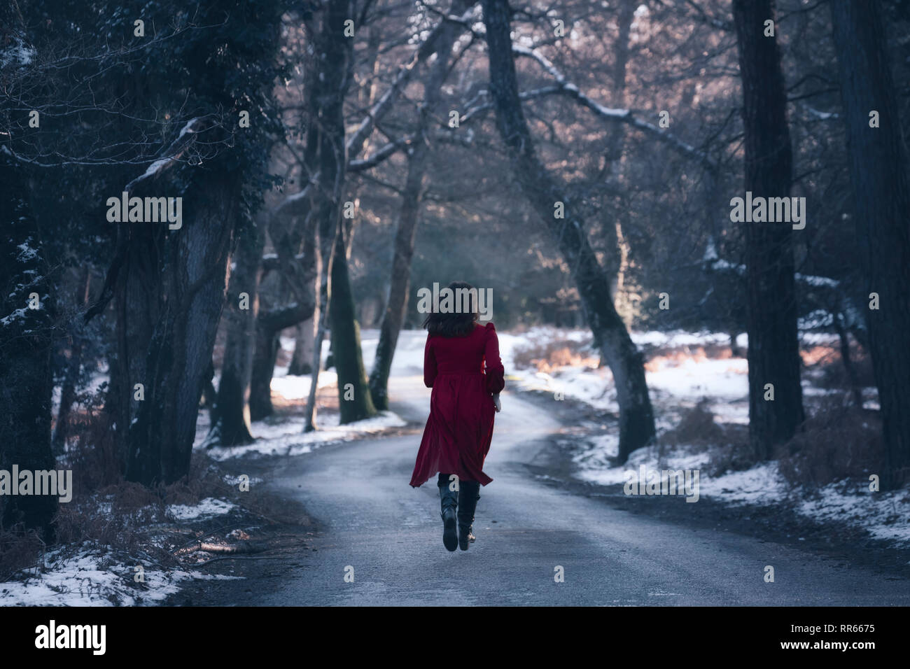 a young woman in a red dress on a small street in the middle of a wintery forest with snow Stock Photo