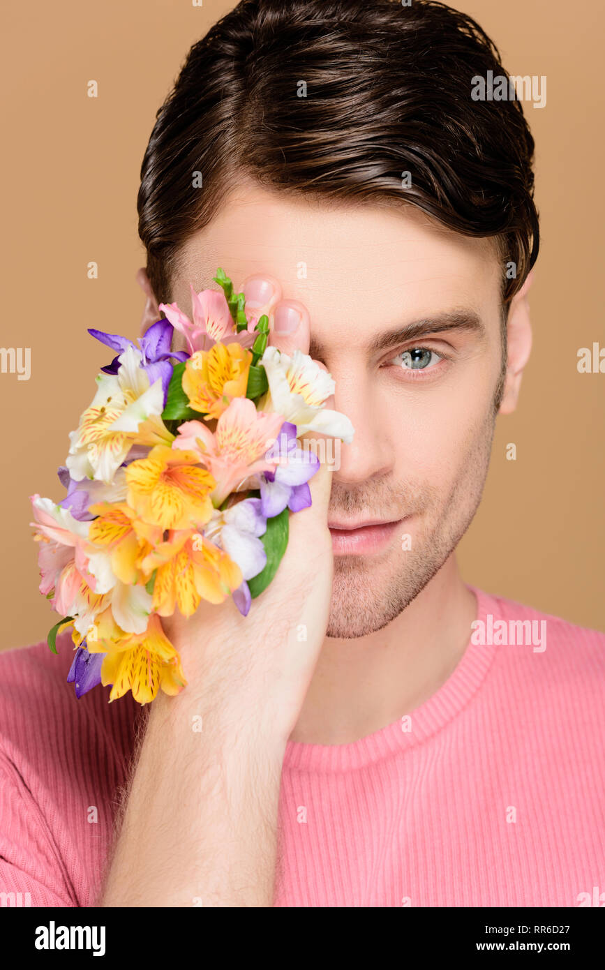 cheerful man covering one eye with flowers on hand isolated on beige ...