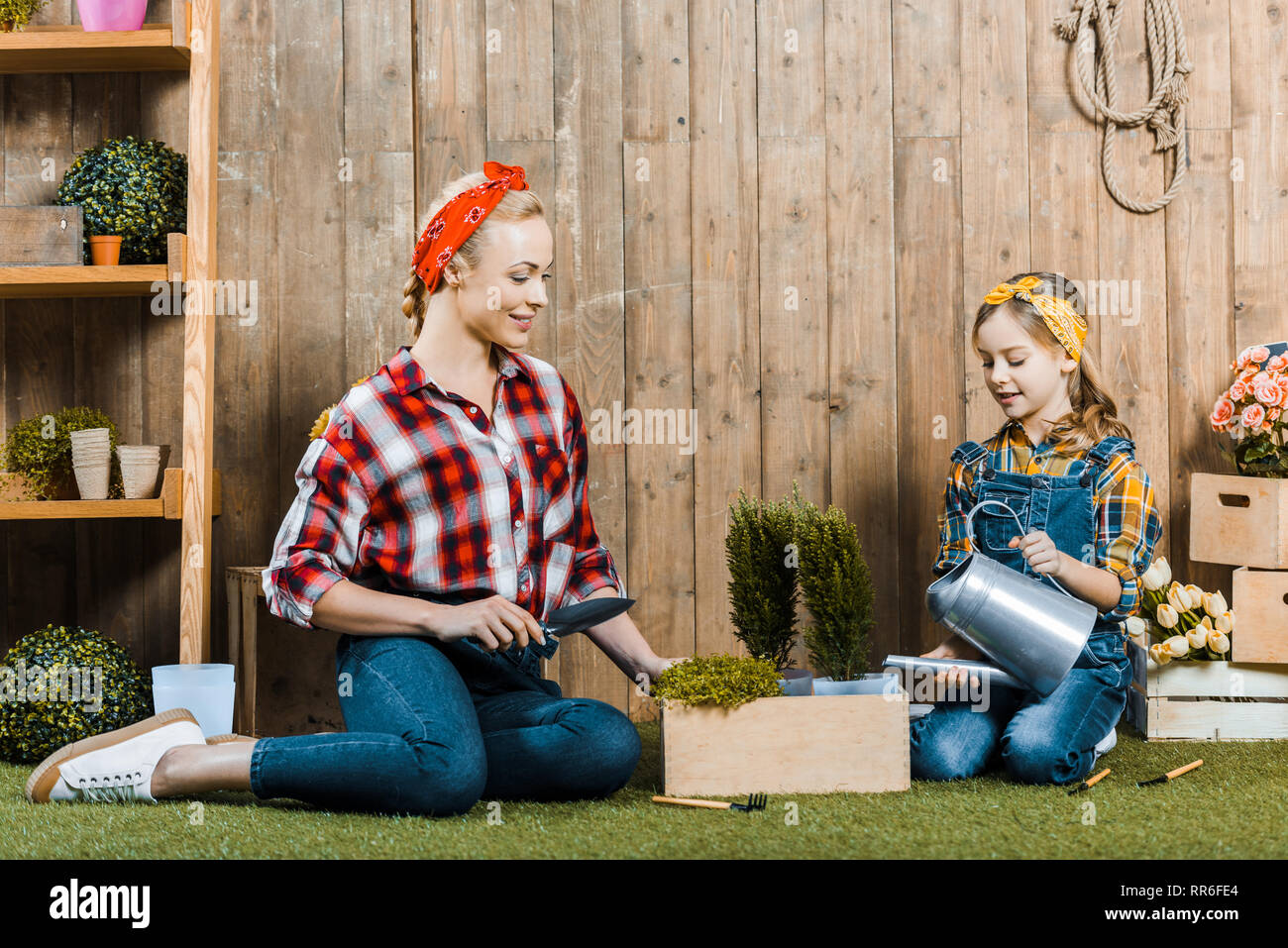 mother holding shovel near daughter watering plant while sitting on grass near wooden fence Stock Photo