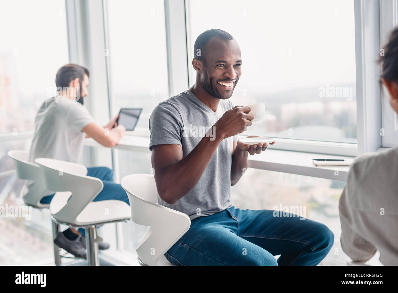 Portrait of multicultural employees dressed in casual wear, laughing while enjoying coffee break during working process in modern office interior, behind glass wall Stock Photo