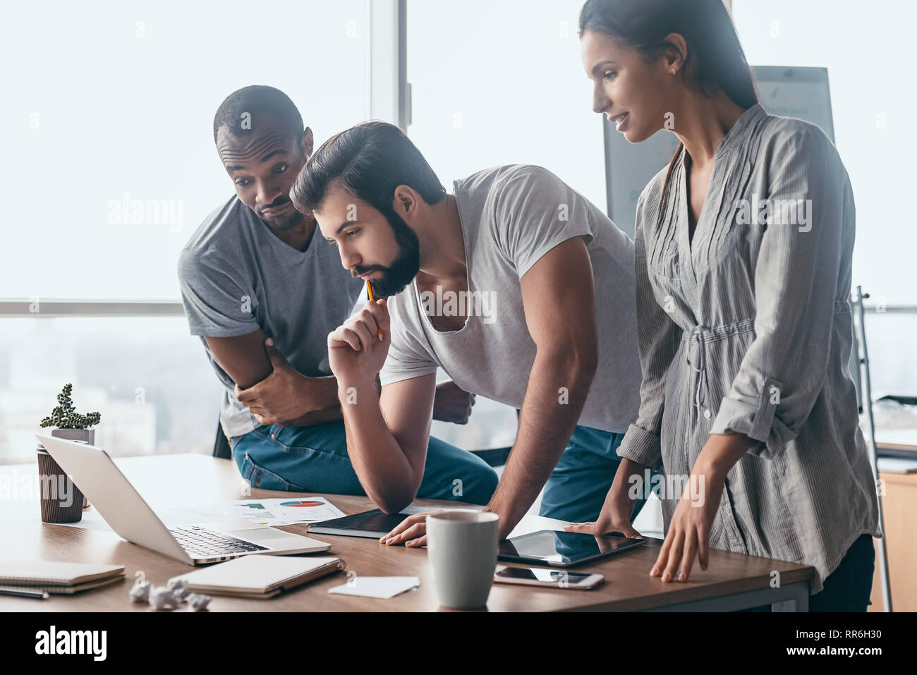 Group of attractive multiracial businesspeople working together in creative office while standing near the wooden desk with a laptop on it, friendly multi-ethnic business team looking on the screen, discussing business issues at meeting Stock Photo