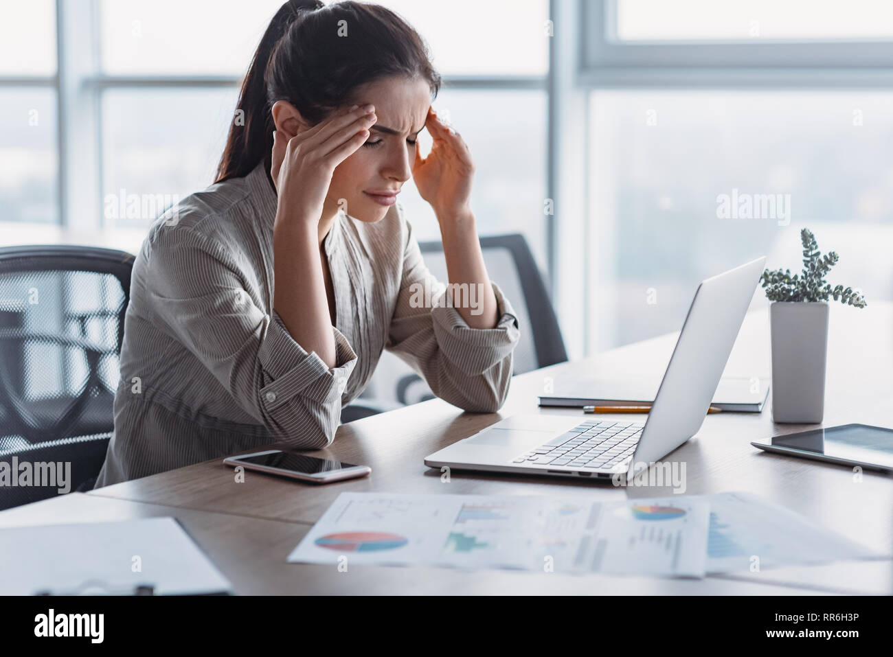 Young dark-haired businesswoman wearing striped shirt is sitting in her office, working on laptop, massaging temples to forget about constant headaches, about noisy office giving a migraine. She is trying to relieve stress and chronic pain Stock Photo