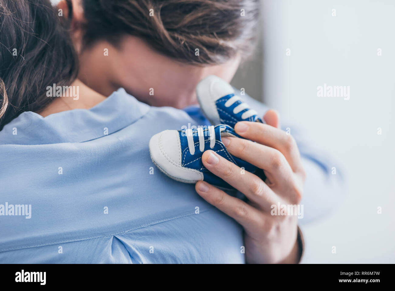 cropped view of man hugging woman and holding blue baby shoes in room, grieving disorder concept Stock Photo